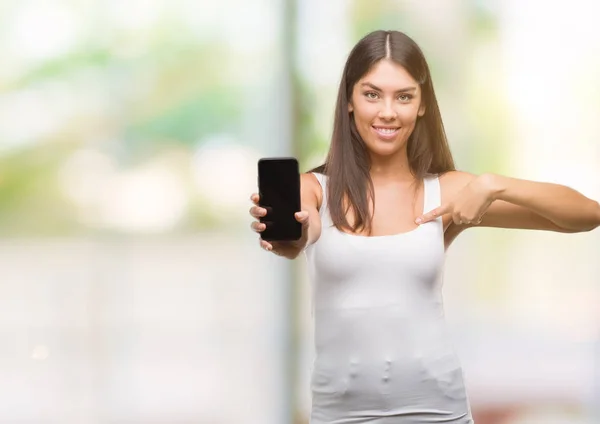 Young beautiful hispanic showing smartphone with surprise face pointing finger to himself