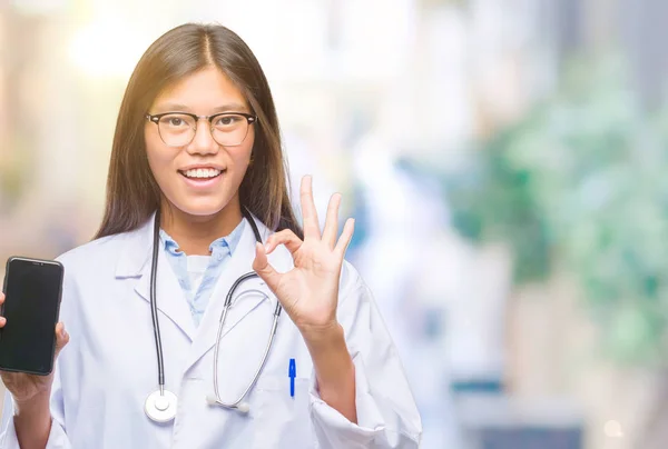 年轻的亚洲医生妇女持有智能手机在孤立的背景下做 标志与手指 优秀的符号 — 图库照片