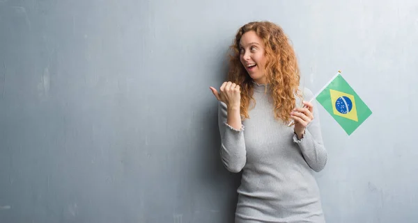 年轻的红发女人在灰色的垃圾墙举行国旗的巴西指向和显示与拇指高兴脸微笑 — 图库照片