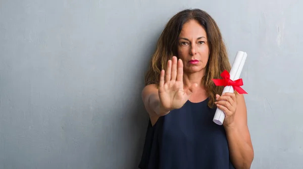 Middle age hispanic woman standing over grey grunge wall holding diploma with open hand doing stop sign with serious and confident expression, defense gesture