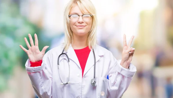 年轻美丽的金发医生妇女穿医疗制服在孤立的背景显示和指向与手指数字七同时微笑自信和快乐 — 图库照片