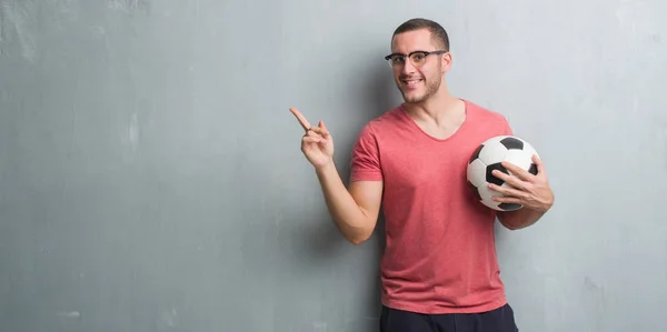 非常に幸せな側の手と指で指すサッカー サッカー ボールを保持している灰色のグランジの壁を越えて若い白人男 — ストック写真