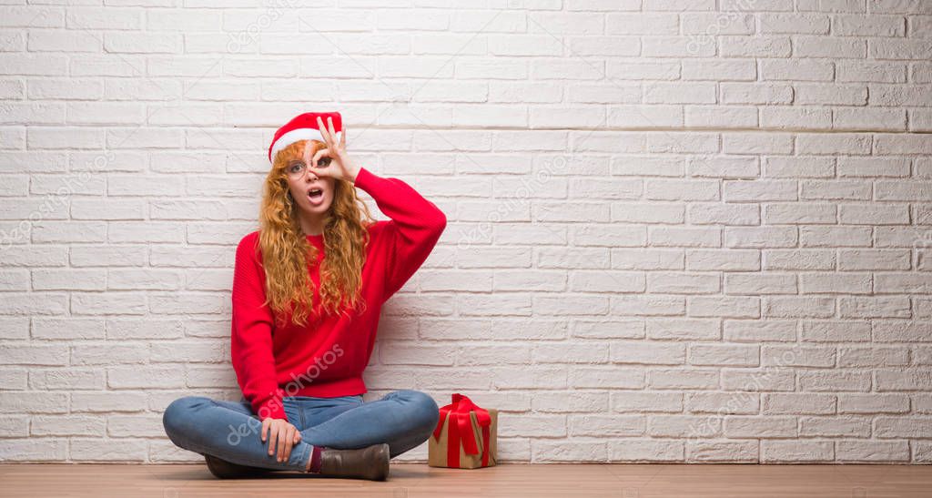 Young redhead woman sitting over brick wall wearing christmas hat doing ok gesture shocked with surprised face, eye looking through fingers. Unbelieving expression.