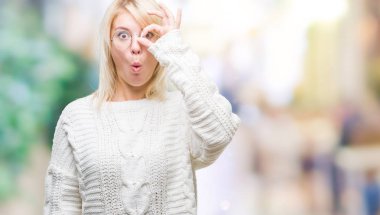 Genç güzel sarışın kadın giyiyor kış kazak ve ok jest sürpriz yüz ile şok yapıyor izole arka plan üzerinde gözlük göz parmaklarının arasından arıyorum. İmansız ifade.