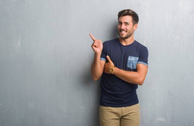 Kameraya bakarak yan el ve parmak ile işaret eden yakışıklı genç gri grunge duvarın yüzünde kocaman bir gülümseme ile üzerinden.