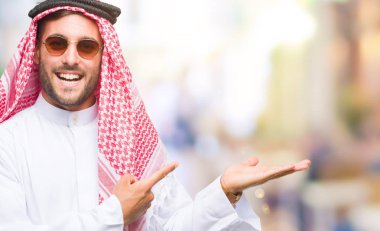 Keffiyeh hayret ve el ile sunulması ve parmak ile işaret ederken kameraya gülümseyen izole arka plan üzerinde giyen genç yakışıklı adam.