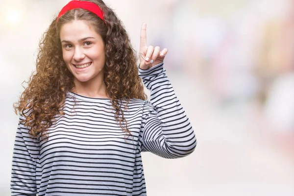 美しいブルネットの巻き毛を着た少女が分離背景表示と自信を持って 幸せな笑みを浮かべて 指番号のいずれかで上向きにセーターをストライプします — ストック写真