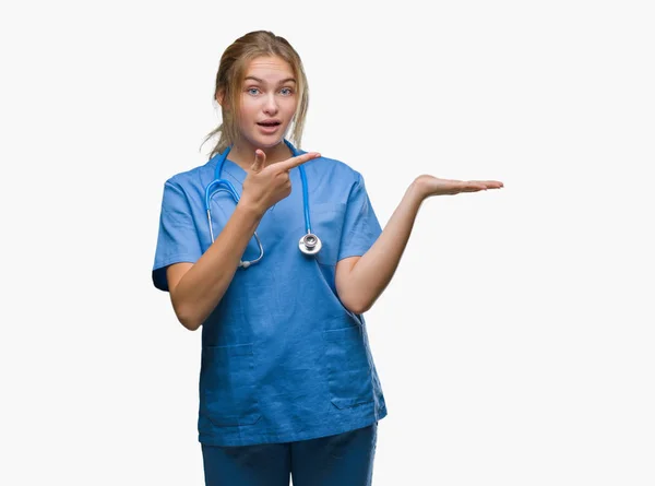 驚くし 手とを提示 指で指している間カメラに笑顔孤立の背景の上の外科医の制服を着て若い白人医師女性 — ストック写真