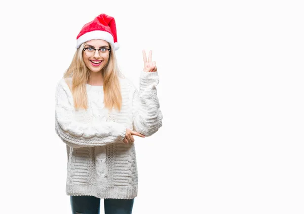 若い美しい金髪女性の勝利のサインを行うカメラでウィンクしている幸せそうな顔を浮かべて隔離された背景にクリスマス帽子をかぶっています — ストック写真
