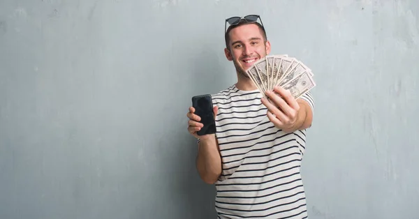 Joven Hombre Caucásico Sobre Pared Grunge Gris Sosteniendo Dólares Teléfono —  Fotos de Stock