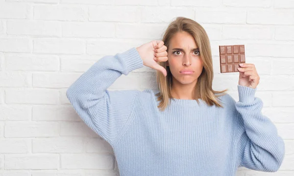 怒った顔 ダウン親指で嫌悪感を示すマイナス記号 拒絶反応の概念とチョコレートバーを白いレンガ壁を食べる以上の美しい若い女性 — ストック写真
