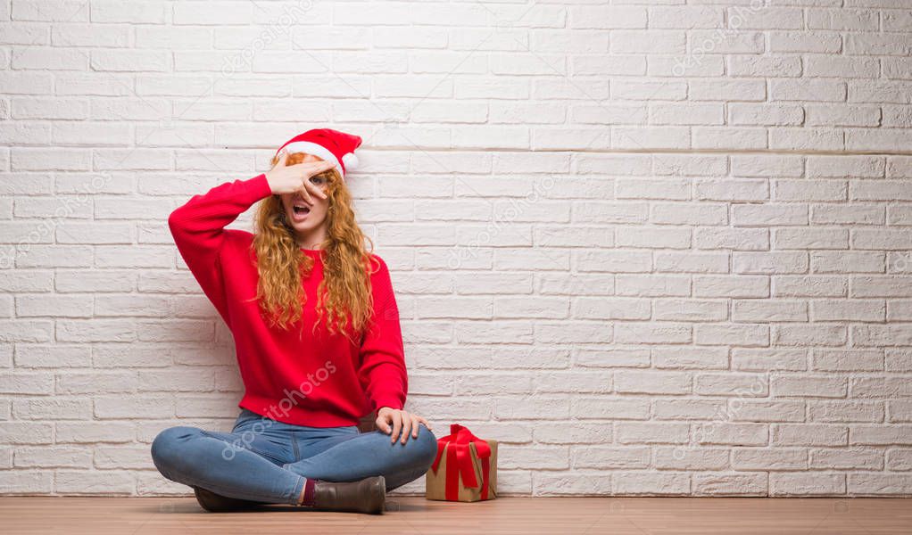 Young redhead woman sitting over brick wall wearing christmas hat peeking in shock covering face and eyes with hand, looking through fingers with embarrassed expression.