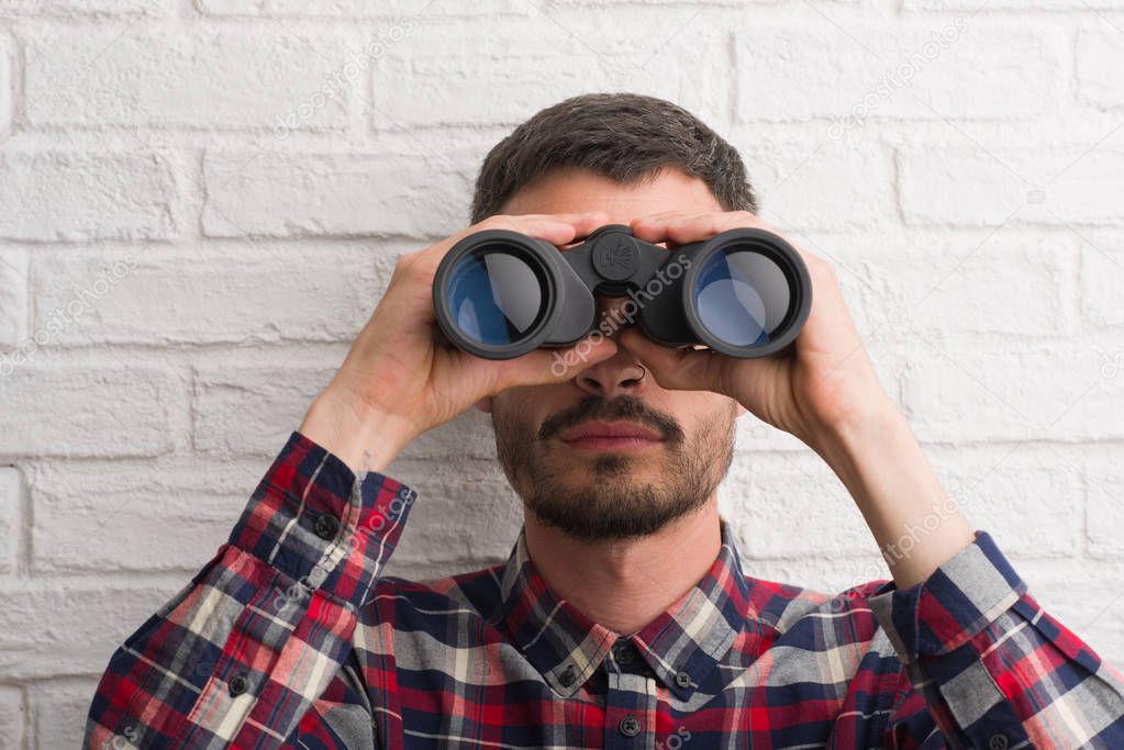 Young adult man over brick wall looking through binoculars with a confident expression on smart face thinking serious
