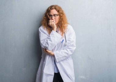 Genç redhead doktor kadın gri grunge duvar stresli ve gergin elle çivi ısırma ağzına bakarak bir palto giyiyor. Anksiyete sorunu.