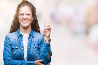 Güzel genç esmer kıvırcık saçlı kız mutlu yüz zafer işareti yaparken kameraya göz kırpıyor ile gülümseyen izole arka plan üzerinde gözlük takıyorum. İki numaralı.