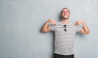 Yüzünde gülümseme ile kendinden emin görünüyorsun, kendini gururlu ve mutlu parmakları ile işaret gri grunge duvarın üzerinden beyaz delikanlı.