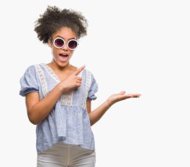 Genç afro Amerikalı kadın hayrete ve el ile sunulması ve parmak ile işaret ederken kameraya gülümseyen izole arka plan üzerinde gözlük.