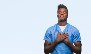 Göğüs kapalı gözler ve yüzünde minnettar jest ile genç Afro-Amerikan Doktor adam cerrah ile Tekdüzen gülümseyen giyen izole arka plan üzerinde eller. Sağlık kavramı.