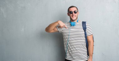 Genç beyaz öğrenci adam giyen kulaklık ve sırt çantası sürpriz ile gri grunge duvarın üzerinden yüz kendine işaret parmağı