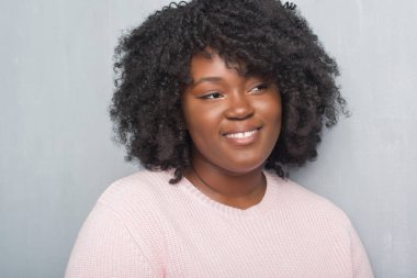 Genç Afro-Amerikan artı boyutu kadın grunge duvar giyen kış kazak uzak tarafına gülümseme ile yüzünde, doğal ifade seyir gri. Kendine güvenen gülüyor.