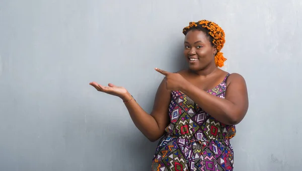 オレンジ三つ編み驚くし 手とを提示 指で指している間カメラに笑顔を身に着けている灰色のグランジの壁を越えて若いアフリカ系アメリカ人女性 — ストック写真