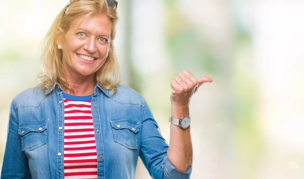 Middle age blonde woman over isolated background smiling with happy face looking and pointing to the side with thumb up.