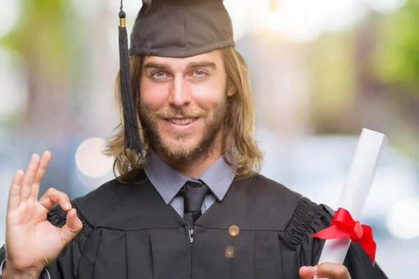ハンサムな若い優秀なシンボルの指で サインをして孤立の背景上の学位を保持する長い髪で男を卒業します — ストック写真