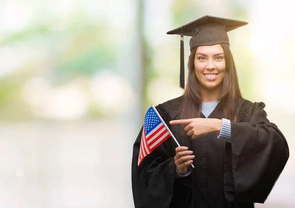 Young hispanic woman wearing graduated uniform holding flag of america very happy pointing with hand and finger