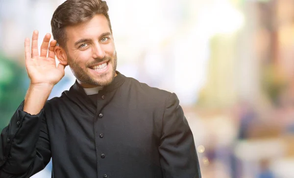 Young catholic christian priest man over isolated background smiling with hand over ear listening an hearing to rumor or gossip. Deafness concept.