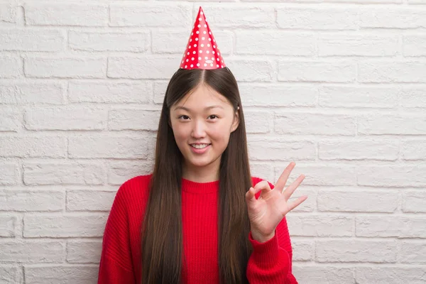 年轻的中国女人在砖墙上戴着生日帽做确定的标志与手指 优秀的象征 — 图库照片