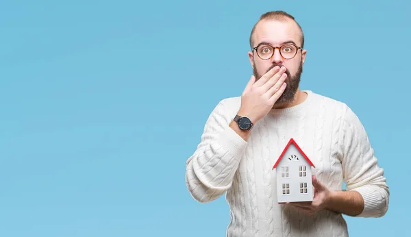 Young caucasian real state agent man holding house isolated background cover mouth with hand shocked with shame for mistake, expression of fear, scared in silence, secret concept