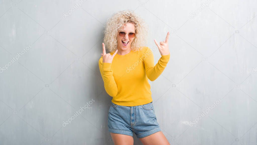 Young blonde woman with curly hair over grunge grey background shouting with crazy expression doing rock symbol with hands up. Music star. Heavy concept.