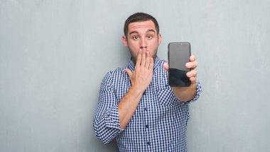 Hata, korku, sessizlik içinde gizli kavramı korkmuş ifade için utanç ile genç beyaz adam eliyle smartphone kapak ağız boş ekran gösterilen gri grunge duvarın üzerinden şok