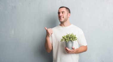 İşaret ve başparmak kadar mutlu yüz gülümseyen tarafı ile gösteren bitki pot tutan gri grunge duvarın üzerinden beyaz delikanlı