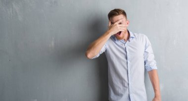 Mahcup ifadeyle parmaklarımın arasından seyir gri grunge duvar yüz ve gözler el ile kapsayan şok bakmak üzerinde genç redhead iş adamı.