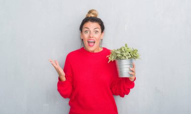 Genç Yetişkin kadın bitki tutan gri grunge duvar yüzünden çok mutlu ve heyecanlı, kazanan ifade büyük gülümseme ile çığlık zaferini kutluyor arsa ve ellerini kaldırdı