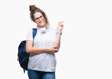 Yüzünde kocaman bir gülümseme ile izole arka plan üzerinde sırt çantası ve kulaklıklar giyen genç esmer öğrenci kız kameraya bakarak yan el ve parmak ile işaret.
