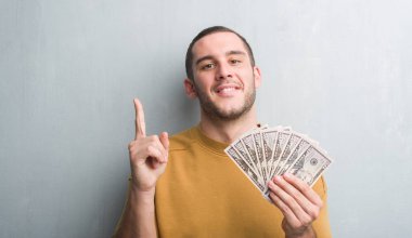 Gri grunge Genç beyaz adam holding dolar bir fikirle sürpriz duvar veya gülen yüz, bir numara ile işaret parmağı soru