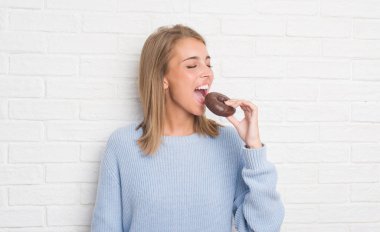 Güzel ve genç birbayan acımak yüz ciddi düşünüyorum üstünde kendine güvenen bir ifade ile çikolatalı çörek yemek beyaz tuğla duvarın üzerinden