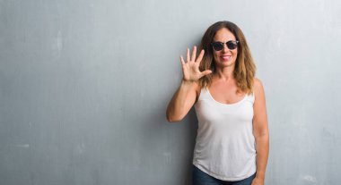 Orta yaş İspanyol kadın gösterilen ve parmakları ile işaret güneş gözlüğü takıyor gri duvar yüzünden kendinden emin ve mutlu gülümseyerek süre beş numara.