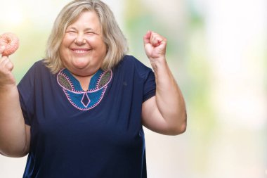 Kıdemli artı boyutu kafkas kadın üzerinde şeker donut yemek büyük gülümseme ile zafer çığlıkları kutluyor ve ellerini kaldırdı arka plan çok mutlu ve heyecanlı, kazanan ifade izole