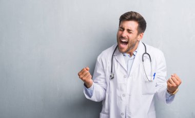 Yakışıklı genç doktor adam gri grunge duvar çok mutlu ve heyecanlı kaldırdı kollarını açarak kazanan jest yapıyor, gülümseyen ve başarı için çığlık. Kutlama kavramı.