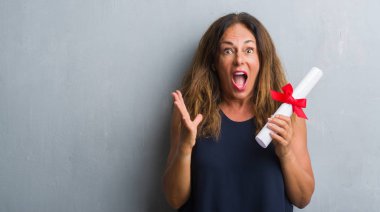 Kocaman bir gülümseme ile çığlık zaferini kutluyor diploması çok mutlu ve heyecanlı, kazanan ifade tutan gri grunge duvarın üzerinden ayakta ve ellerini kaldırdı orta yaş İspanyol kadın