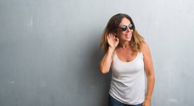 Orta yaş İspanyol kadın gri duvar bir işitme söylenti ya da dedikodu için dinleme kulak üzerinde el ile gülümseyen güneş gözlüğü takıyor. Sağırlık kavramı.