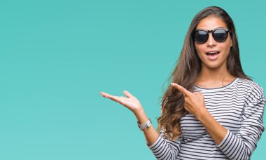 Genç güzel Arap kadın hayrete ve el ile sunulması ve parmak ile işaret ederken kameraya gülümseyen izole arka plan üzerinde güneş gözlüğü takıyor.