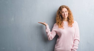 Gri grunge duvar neşeli sunulması ve kameraya bakarak avucunun ile işaret gülümseyen pembe kazak giyen genç Kızıl saçlı kadın.