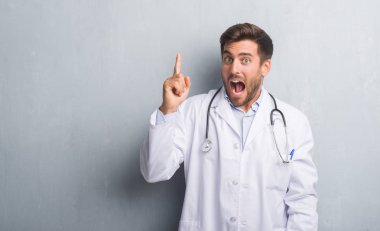 Yakışıklı genç doktor adam gri grunge başarılı fikirle işaret parmağı duvar. Çıktıktan ve mutlu. Bir numara.