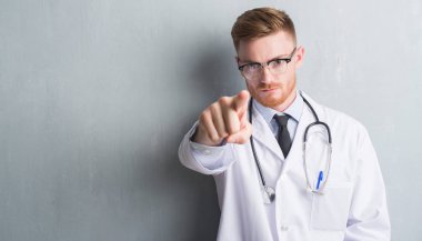 Genç redhead Doktor adam kamera ve size parmak ile işaret gri grunge duvarın üzerinden el işareti, pozitif ve kendinden emin bir jest ön