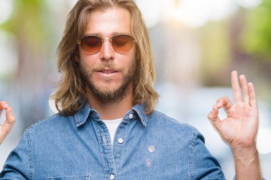 İzole arka plan üzerinde güneş gözlüğü takmış uzun saçlı yakışıklı delikanlı rahatla ve gülümseyen gözlerle kapalı parmak jestle meditasyon yapıyor. Yoga kavramı.