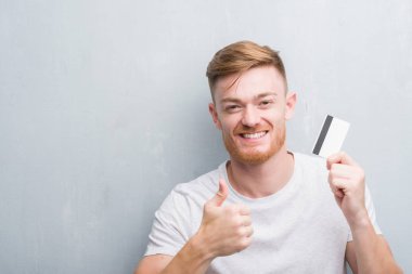 Kredi kartı ile mutlu büyük holding gri grunge duvarın üzerinden genç Kızıl saçlı adam gülümseme ok işareti, başparmak ile parmak, mükemmel işareti yapıyor